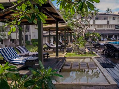 Venkovní hotelová oblast s dlážděným bazénem, lehátky a rostlinami pod modrou oblohou.