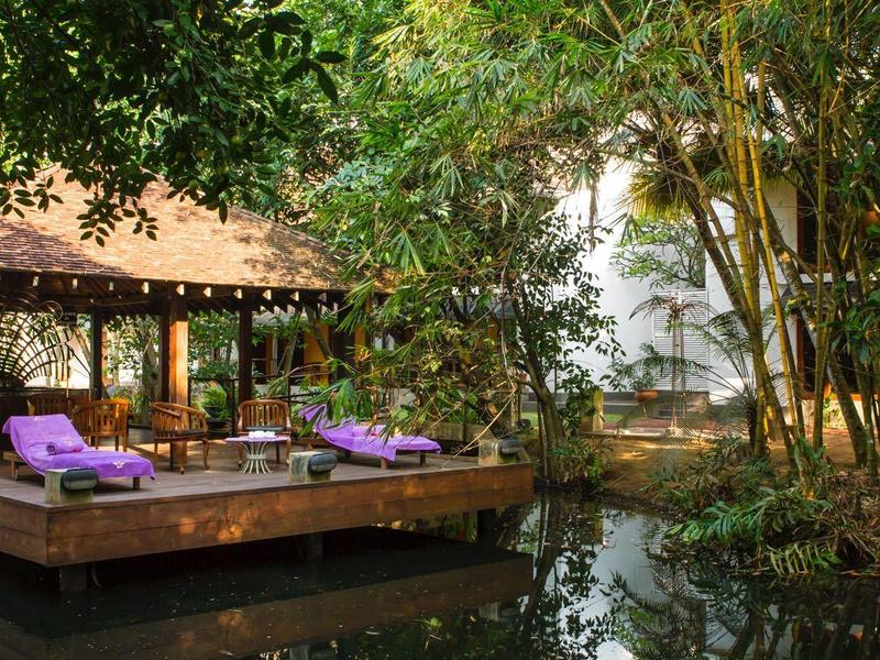 Venkovní lounge s dřevěnou podlahou a fialovými lehátky vedle vody a bujné zeleně.