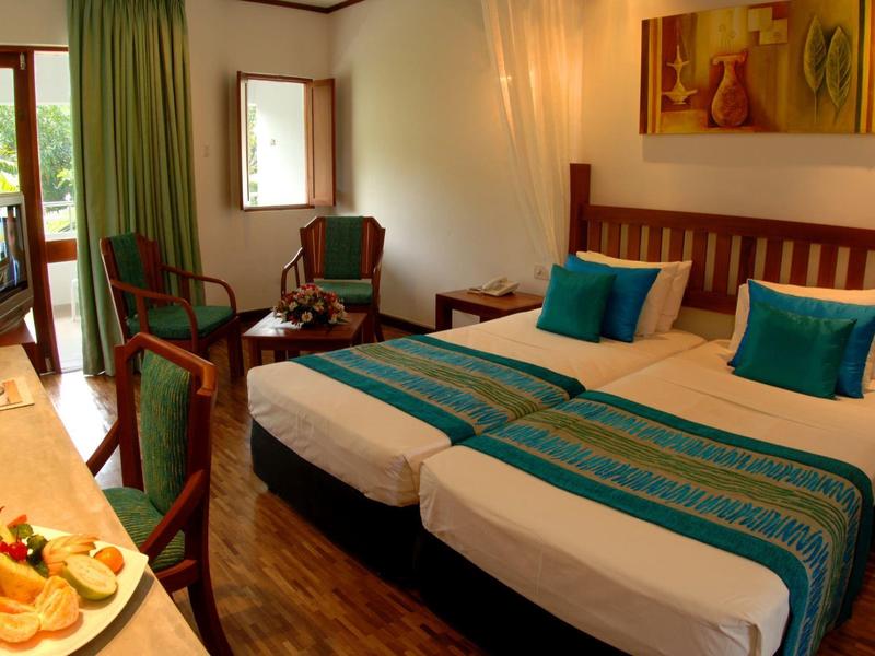 Světý hotelový pokoj se dvěma jednolůžky, modrými polštáři a přístupem na balkon s výhledem na zeleň.