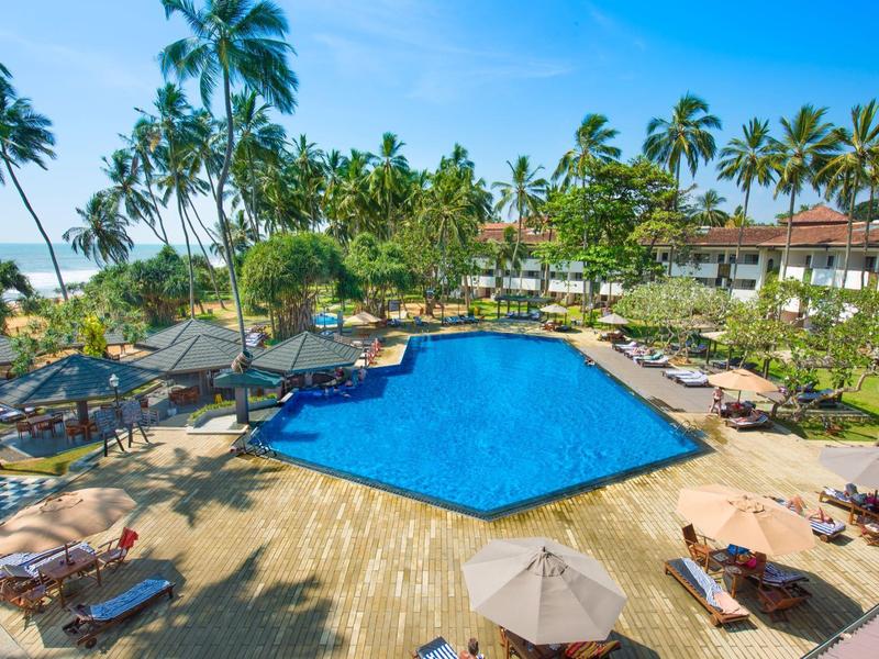 Velký venkovní bazén obklopený lehátky, palmami a hotelovými budovami za slunečného počasí.