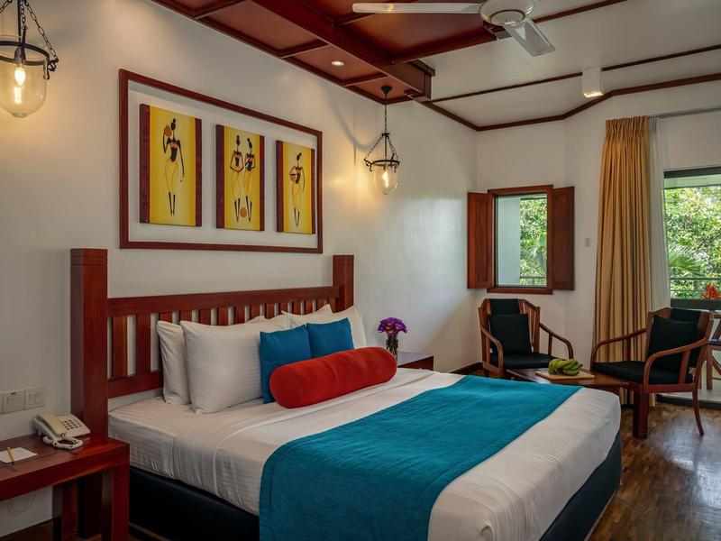 Elegantní hotelový pokoj s manželskou postelí, dřevěným nábytkem a výhledem na zelené stromy oknem.