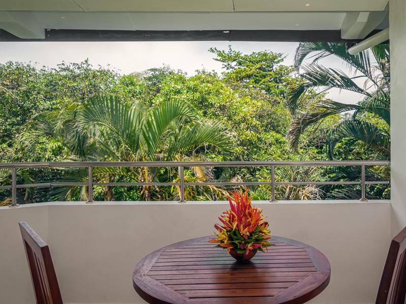 Balkon s malým dřevěným stolem, dvěma židlemi a květinovou dekorací na pozadí zelené džungle.