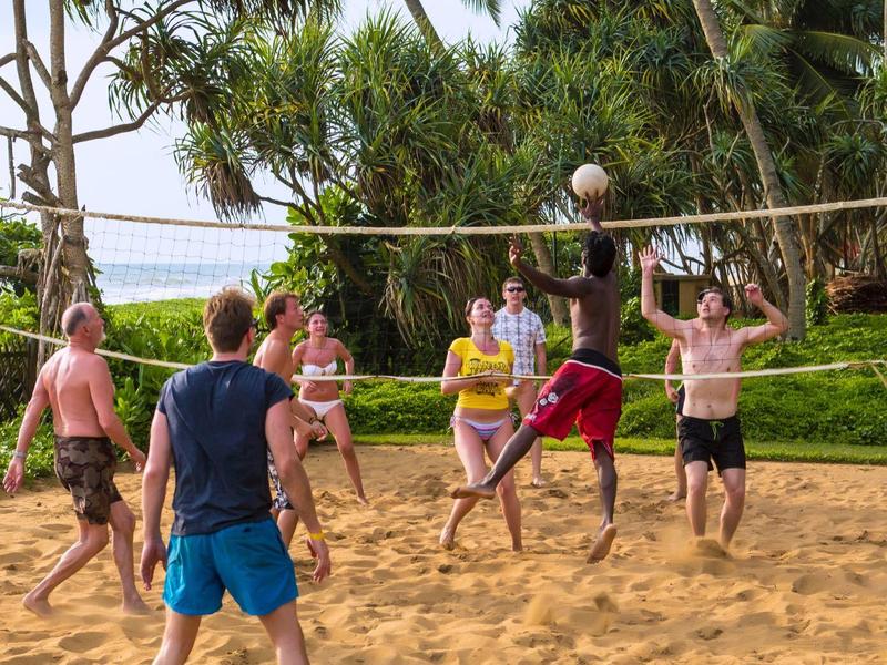 Skupina mladých lidí hrající beach volejbal na pískovém hřišti obklopeném palmami.