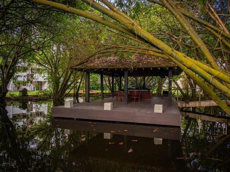 Dřevěná zastřešená plošina na klidné tůni obklopená hustou zelení a bambusem.