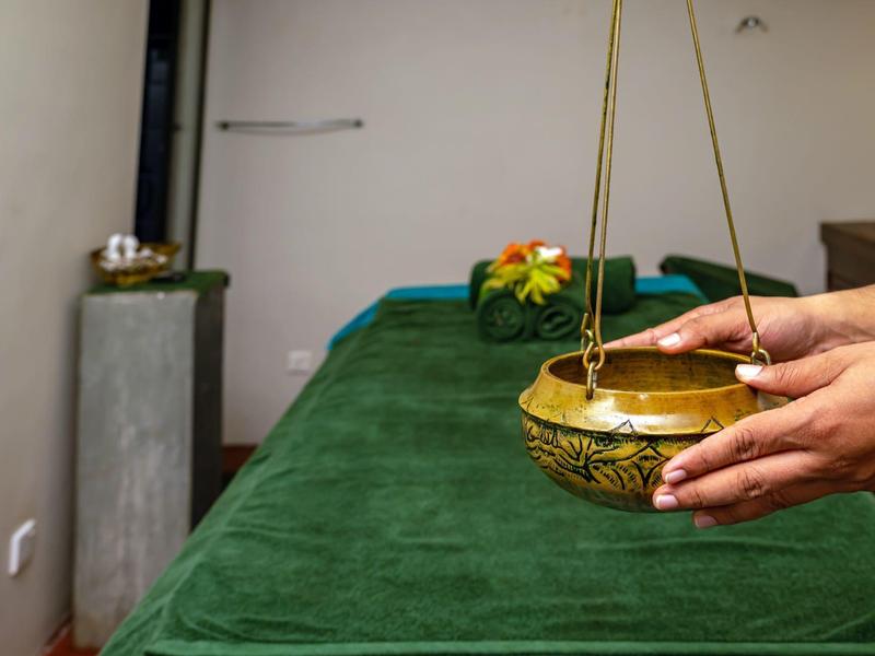 Osoba drží zavěšenou aromatickou misku nad zeleným masážním lehátkem v relaxační místnosti.