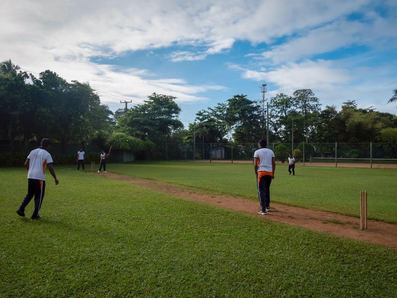 Lidé hrají kriket na zeleném hřišti pod zataženou oblohou.