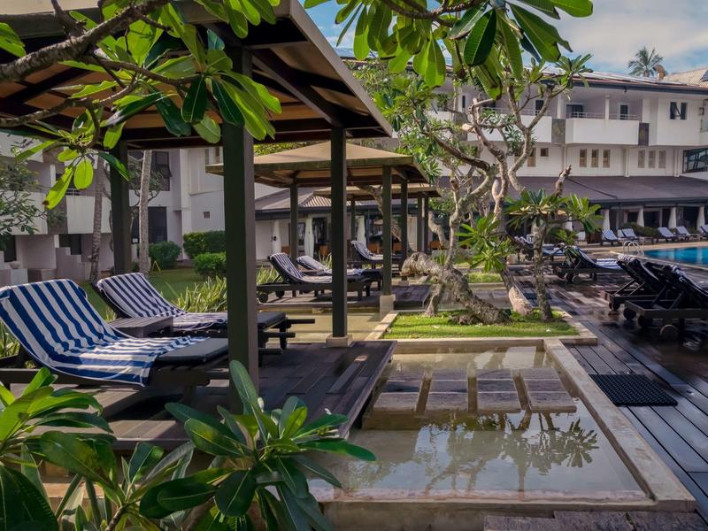Venkovní hotelová oblast s dlážděným bazénem, lehátky a rostlinami pod modrou oblohou.