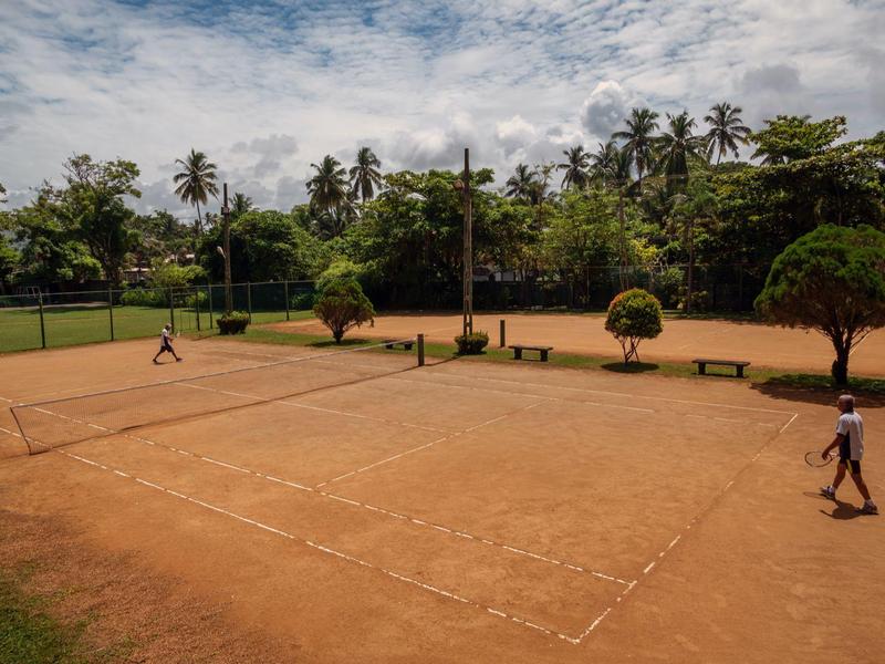 Dva lidé hrají tenis na antukovém kurtu obklopeném palmami a zelení.