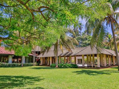 Großes tropisches Haus mit Strohdach, umgeben von grünen Bäumen und weitläufigem Rasen.