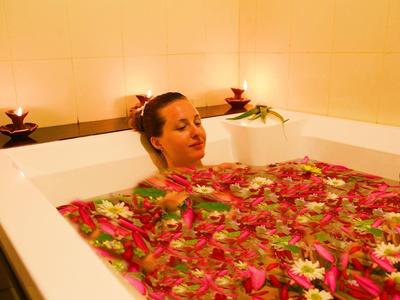 Frau entspannt in Badewanne mit Wasser und vielen rosa Rosenblättern, warmes Licht im Raum.