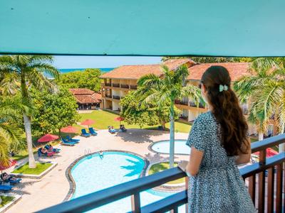 Frau schaut von Balkon auf Pool, Palmen und Gebäude in sonniger tropischer Hotelanlage.