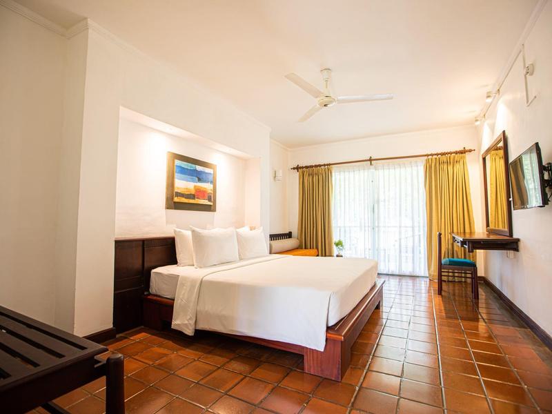 Helles Schlafzimmer mit großem Bett, Fliesenboden, Wandbild, Deckenventilator und Balkon mit Vorhängen.