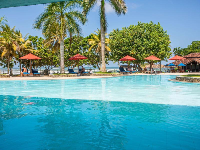 Großer blauer Pool mit Palmen, Sonnenschirmen und klarem Himmel im Hintergrund.