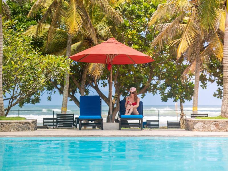 Pool mit blauem Wasser, zwei blauen Liegestühlen, rotem Sonnenschirm und Palmen am Strand.