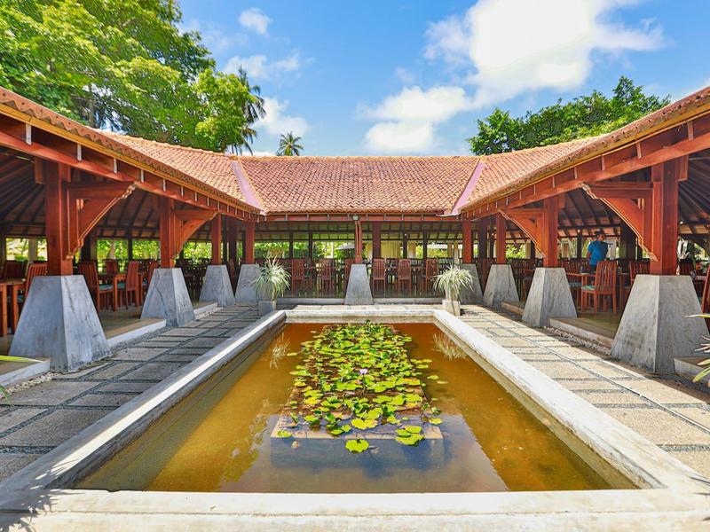 Überdachter Holzpavillon mit Teich und Seerosen, umgeben von grünen Bäumen und blauem Himmel.