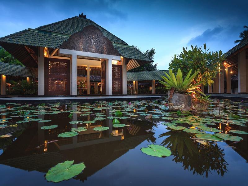 Traditionelles Haus mit grünem Dach neben Teich mit Seerosen und Pflanzen am Ufer bei Abenddämmerung.