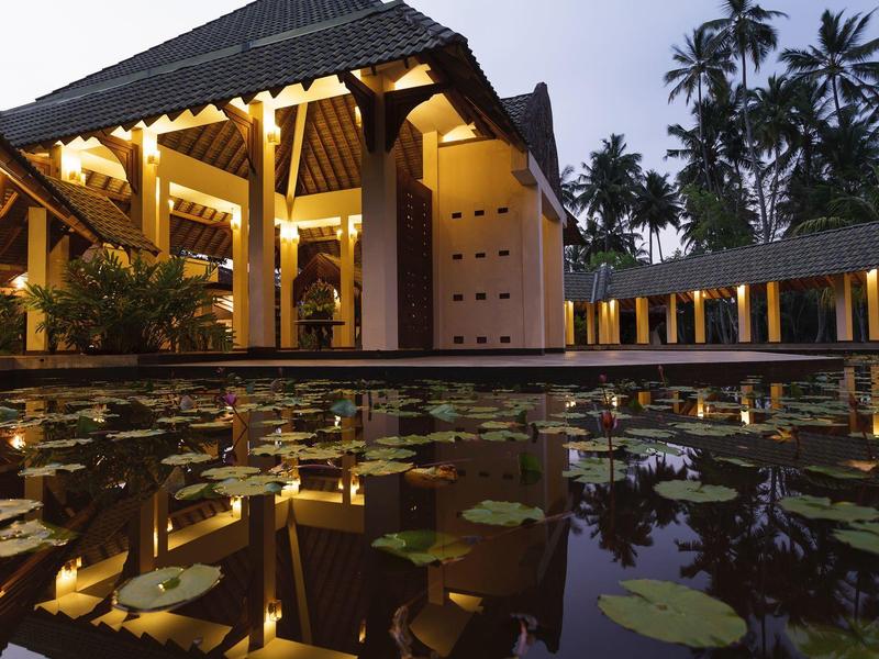 Ein beleuchtetes Haus mit offenem Innenraum neben einem Teich mit Seerosenblättern bei Dämmerung.