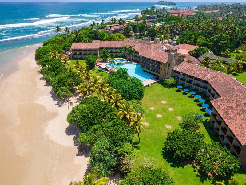 Große Hotelanlage mit Pool, umgeben von Palmen, direkt an einem weißen Sandstrand am blauen Meer.