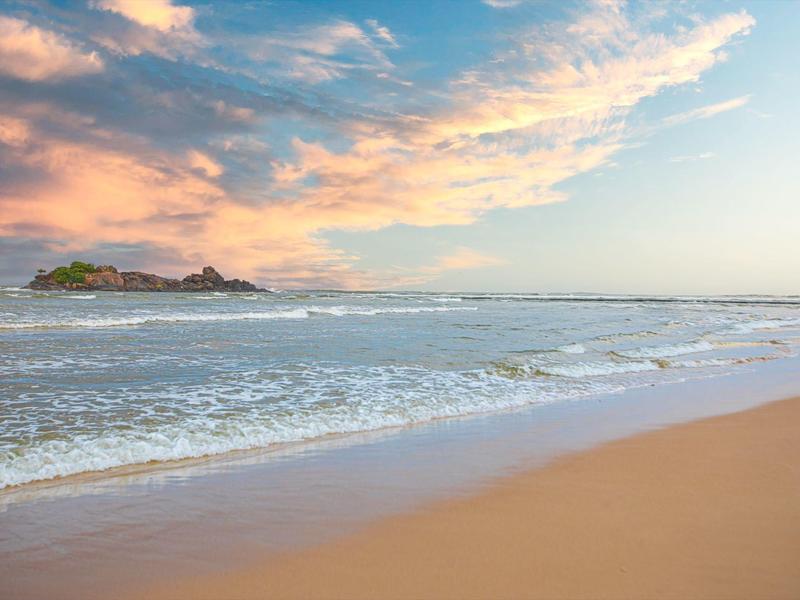 Sandstrand mit sanften Wellen, weiter Horizont und bunter Himmel mit Wolken bei Sonnenuntergang.