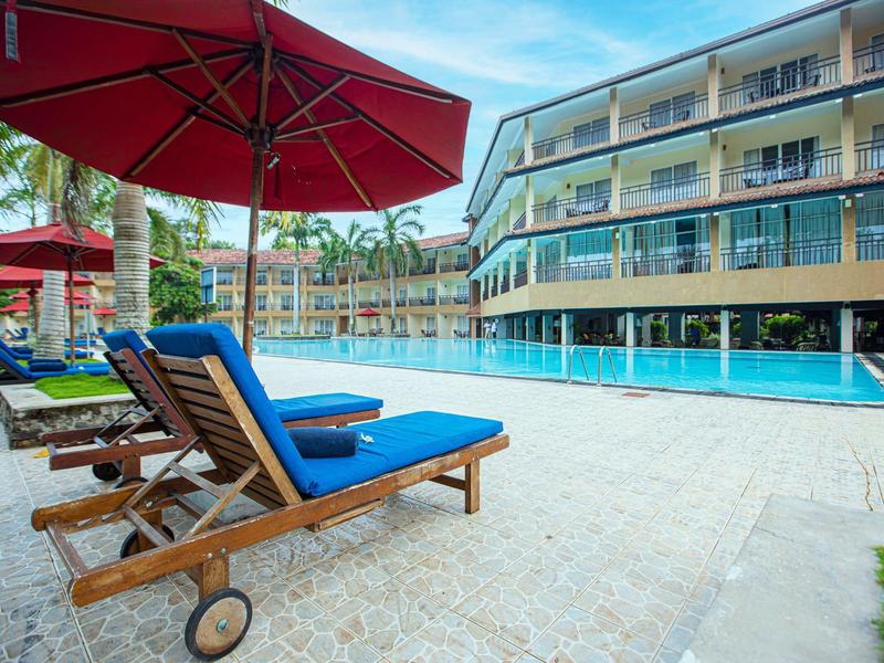 Ein Poolbereich mit blauen Liegestühlen, roten Sonnenschirmen und modernem Hotelgebäude im Hintergrund.