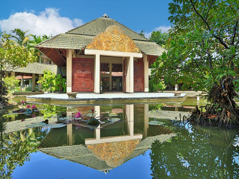 Ein traditionelles Haus am Teich mit Spiegelung, umgeben von Bäumen und blauem Himmel.