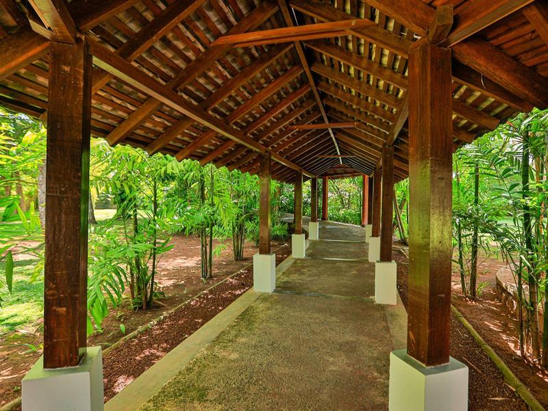 Ein überdachter Holzweg durch einen grünen, bewaldeten Garten mit Steinboden und Säulen.