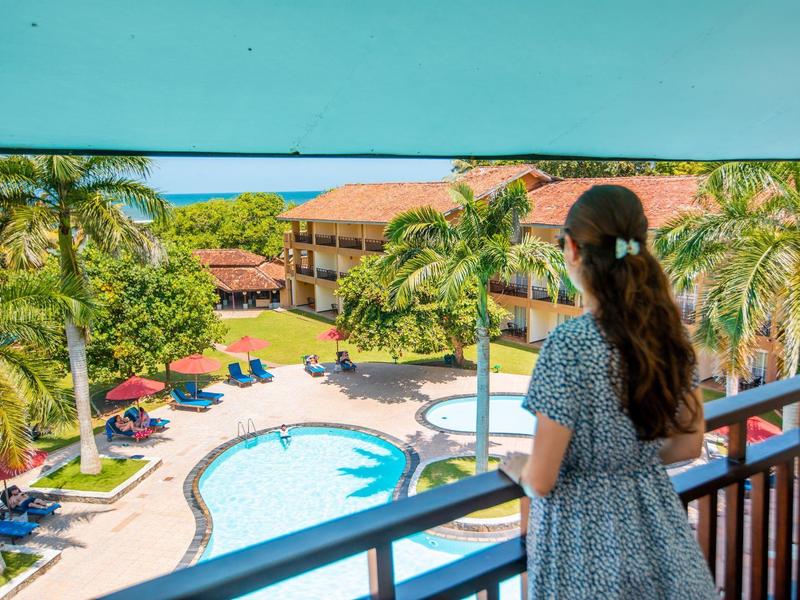 Frau schaut von Balkon auf Pool, Palmen und Gebäude in sonniger tropischer Hotelanlage.
