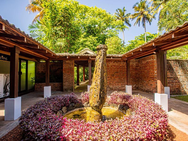 Innenhof mit Brunnen, Rundbeet mit roten Pflanzen, rot-braune Mauern und grünem Baum hinter.
