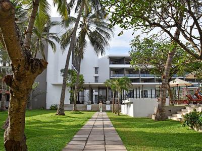 Path leads through tropical garden to a modern multi-story hotel building with balconies.