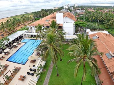 View of a hotel area with pool, sun loungers, palm trees, and green lawns by the sea.