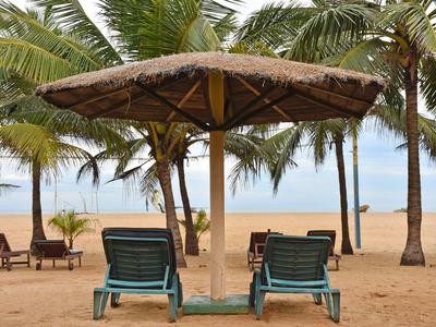 Two lounge chairs under a thatched roof on a beach with palm trees in the background.