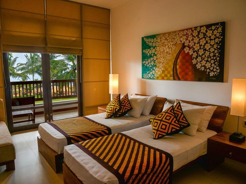 Bright hotel room with double bed, decorative pillows, and balcony overlooking palm trees.