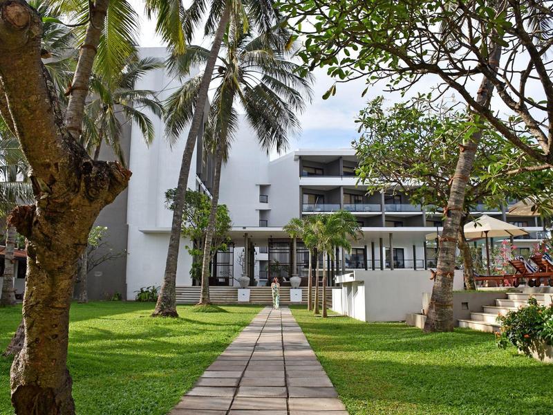 Path leads through tropical garden to a modern multi-story hotel building with balconies.
