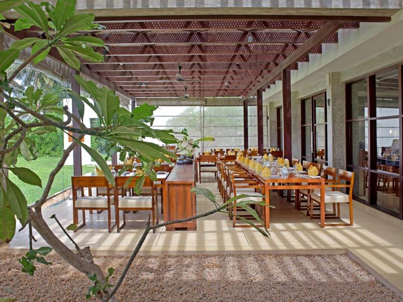 Covered outdoor dining area with tables and chairs next to a building, plants visible.