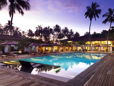Hotelpool mit Holzdeck und beleuchteter Lounge bei Sonnenuntergang, umgeben von Palmen.