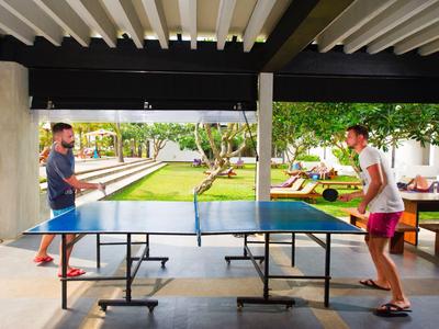 Zwei Männer spielen Tischtennis unter einem überdachten Bereich neben einer grünen Gartenanlage.