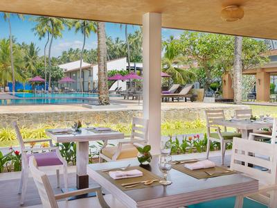 Überdachte Terrasse mit Tischen und Stühlen, Blick auf Pool und Palmen in Hotelanlage.