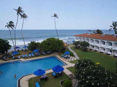 Hotel mit Pool, blauen Sonnenschirmen und Blick auf das Meer bei klarem Himmel.