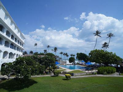 Hotel mit mehrstöckigem Gebäude, Garten, Pool und Palmen unter blauem Himmel.