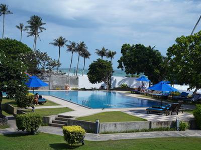 Pool mit Sonnenliegen, blauen Sonnenschirmen und Palmen vor dem Meer unter bewölktem Himmel.