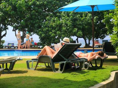 Zwei Frauen entspannen auf Liegestühlen neben einem Pool mit Bäumen und blauem Sonnenschirm.