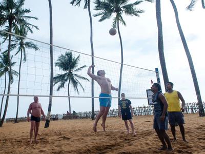 Menschen spielen Beachvolleyball auf einem Sandplatz mit Palmen im Hintergrund.