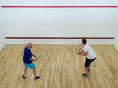 Zwei Männer spielen Squash auf einem hellen Holzboden in einem Squashcourt mit weißen Wänden.