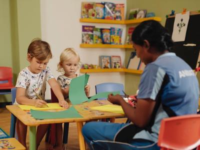 Children crafting with help from a caregiver at a table in a colorful room.