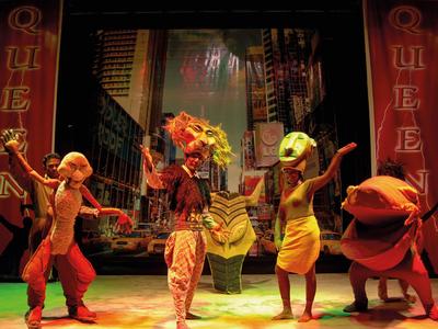 Vier farbenfrohe Darsteller in Kostümen auf Theaterbühne vor großem Stadt-Hintergrund