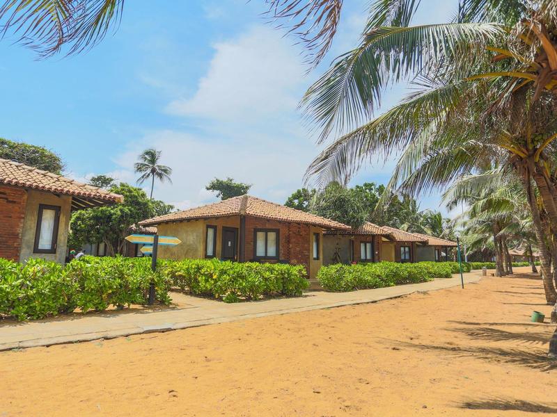 Row of small holiday bungalows with palm trees along a sunny beach path.