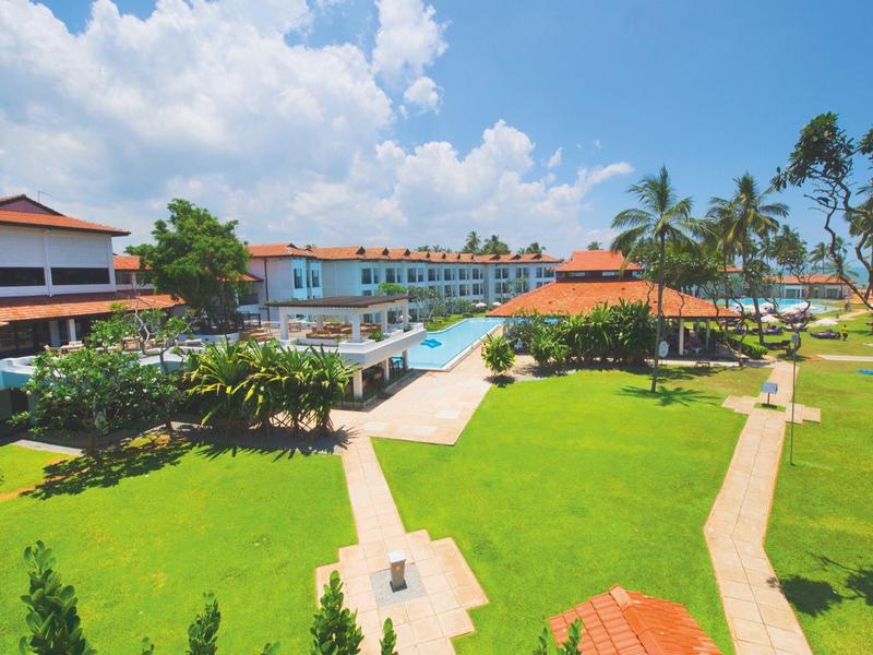 Large garden with green lawns and swimming pool beside hotel buildings under blue sky