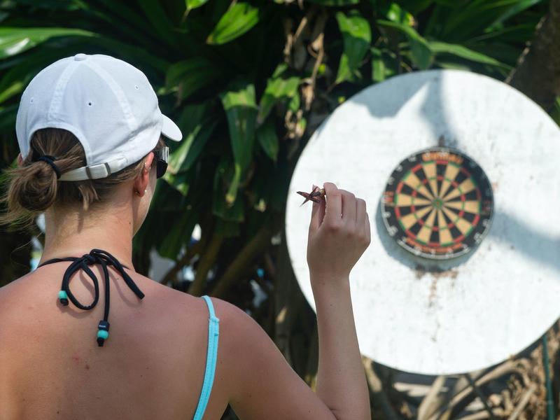 Woman wearing white cap playing darts outdoors in front of a leafy wall.