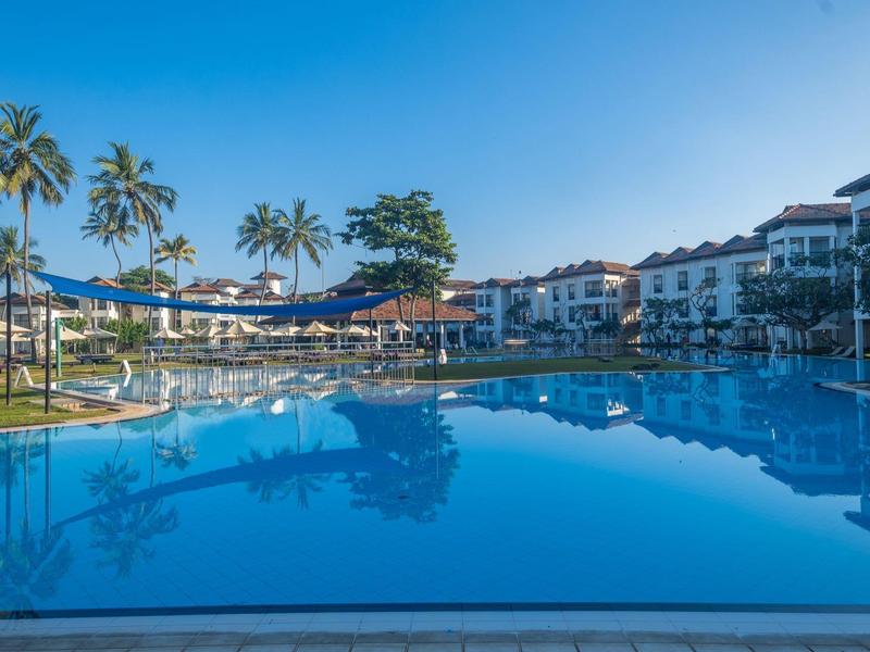 Large swimming pool with clear water in front of hotel buildings and palm trees under a sunny sky.