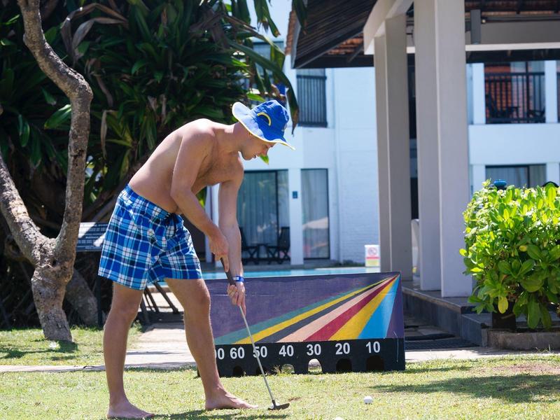 Man in blue swim trunks and hat plays mini golf on grass beside a tree and building.